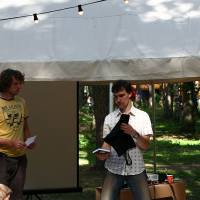 Three individuals are in front of a tent at Positivus 2008. One person on the left is holding a paper and looking at a person in a white shirt who is speaking and displaying a black object. Another person on the right, wrapped in a patterned shawl, looks on. A string of lights decorates the tent.