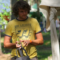 A man with curly hair, wearing a yellow "Big League" T-shirt, holds a black tote bag with a Fairtrade logo. He stands outside near a large tent at the 2008 Positivus festival, surrounded by greenery and trees, writing in a small notebook or adjusting an item in his hands. Sunlight filters through the trees.
