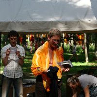 A person wearing glasses and an orange towel around their shoulders stands under a canopy at the Positivus 2008 festival, holding a small black box and other items. Another person nearby holds a sheet of paper while a third person is crouched near a tree. Trees and the white canopy are visible in the background.