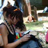 A person with brown hair, wearing an orange headband and sunglasses on their head, sits on grass reading a booklet during Positivus 2008. Clad in a black tank top, multiple wristbands, and a necklace with a skull pattern, they exude festival vibes. Meanwhile, another person is bent over, rummaging through a box in the background.
