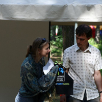 Two people stand outdoors under a white canopy in a wooded area during the Positivus festival. The woman on the left holds a microphone and appears to be speaking while holding a black bag with a blue handle. The man on the right, in an AB 2008 patterned shirt, looks at her attentively.