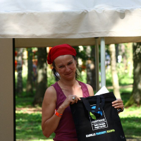 A person wearing a red headscarf and a sleeveless maroon top is standing outdoors under a canopy at the Positivus festival. They are holding a black tote bag with white text that includes "Fairtrade." Trees in the background suggest a park setting. The person is smiling slightly, with one eye squinted.