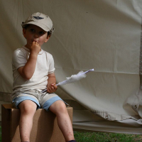 A young child sits on a cardboard box, wearing a beige cap, white t-shirt, light blue shorts, and sandals. They hold cotton candy in one hand with a thoughtful expression. The background features a large white fabric tent at Positivus AB in 2008.