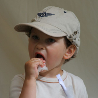 A young child wearing a beige baseball cap with an AB 2008 emblem is eating a piece of white cotton candy from a paper stick. The child, dressed in a white shirt, has a focused expression while enjoying the treat against a simple beige backdrop—bringing back memories of Positivus festival joy.