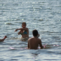 Three people are swimming in the ocean. One person in the center, wearing a bikini, appears to be splashing water playfully towards the two others, who are closer to the foreground and shirtless. The water is shimmering under the sunlight, creating a lively and fun atmosphere reminiscent of Positivus AB 2008.