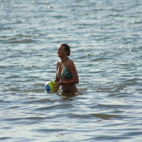 A person stands waist-deep in the water, holding a yellow and blue volleyball, reminiscent of sunny days at Positivus 2008. The individual is wearing a swimsuit and enjoying the serene beach, surrounded by calm waves. The background shimmers with the glistening water surface.