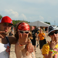 Three young men pose on a beach on a sunny day at Positivus AB 2008. One wears a red cap and white tank top, the middle person sports sunglasses, a red bandanna, and gestures with an open hand, and the third wears a patterned cap and colorful shirt, gesturing with two fingers. A tent and crowd are visible in the background.