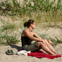 A woman sits on a red towel at a sandy beach during the Positivus festival, surrounded by green plants and grass. She is looking to the side, wearing a black tank top, sunglasses, and green shorts. Beside her are a pair of green sandals and a white item, possibly a towel or clothing.