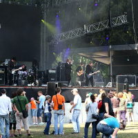 A band performs on a large outdoor stage at Positivus, with various instruments under bright lights. Several audience members stand and watch, some wearing casual clothes, reminiscent of AB 2008. The stage setup includes amplifiers, speakers, and scaffolding. Trees in the background suggest a park setting.