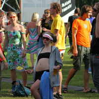 A group of people enjoying an outdoor event at Positivus. In the foreground, a person in a gray hoodie, wearing sunglasses and a sun visor, is energetically dancing while holding a beer. Others are standing or walking around, talking and laughing. A large banner reading "AB 2008" and various colorful outfits are visible in the background.