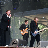 At Positivus AB 2008, a band performs on an outdoor stage. One member stands at the microphone singing, another plays bass guitar, and the third plays electric guitar. All are dressed in black suits. Stage equipment, including large speakers and amps, surround them, with trees visible in the background.