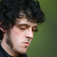 A man with curly dark hair, sideburns, and a pensive expression is shown looking down. Wearing a black jacket, red tie, and striped shirt, his attire suggests it might be from 2008. The background is blurred with a greenish tone, possibly indicating an outdoor setting at Positivus. The focus is on his face and upper body.