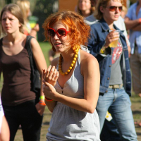 A young woman with bright orange hair, wearing red sunglasses, a grey dress, and a yellow beaded necklace, is walking outside among a crowd at Positivus 2008. Other people in casual attire are in the background, with some wearing jeans and T-shirts. Green and white tents are visible in the distance.