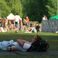 A boy wearing sunglasses lies on the grass next to a soccer ball and black backpack. In the background, people gather, sit, and socialize near white tents and a metal barrier at an outdoor event reminiscent of Positivus. Trees and greenery surround the open space, giving the scene a relaxed, summery atmosphere.