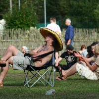 A man wearing a large sombrero and shorts reclines in a foldable chair on a grassy field during the 2008 Positivus festival, enjoying the lively outdoor event with other casually dressed attendees conversing in the background. A wooden fence and trees frame the distant AB landscape.