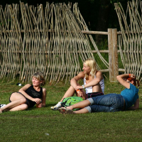 Three people are sitting on grass near a wooden fence. Two are seated: one on the left in a black top and the other in the middle with a white top and green bag. A third person, wearing a blue top, is lying down on the right. The background channels Positivus 2008 vibes with its unique fence made of vertically interwoven branches.