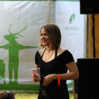 A smiling woman with shoulder-length brown hair wearing a black dress and an orange wristband holds a clear plastic cup. She stands under a green and white canopy with a shadowed deer emblem at Positivus AB 2008. People seated on the grass and sound equipment are visible in the foreground and background.