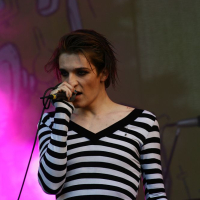 A person with short wet hair is performing on stage at Positivus, holding a microphone close to their mouth. They are wearing a black and white striped, long-sleeved top. The background is illuminated with vibrant purple and pink stage lights, reminiscent of the AB 2008 vibe. The overall atmosphere suggests a lively concert or music performance.