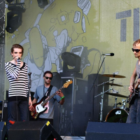 Four musicians perform on an outdoor stage with a yellow backdrop featuring abstract art at Positivus AB 2008. The lead singer, wearing a striped shirt, holds a microphone, while a guitarist in sunglasses and a sleeveless shirt stands to his right. A bassist and drummer are at the back, playing their instruments.