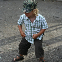 A young child with blonde hair stands on a paved surface, wearing a green camouflage cap, a white and green plaid short-sleeve shirt, dark pants, and open-toed sandals. The child is bending slightly forward with both hands on their knees. A black backpack from Positivus AB 2008 is in the background.