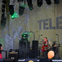 Three musicians perform on a stage with various instruments, including a guitar and bass, under the AB 2008 banner. The backdrop features the "Tele2" logo surrounded by abstract art. Multiple stage lights illuminate the performers while yellow balloons float near the stage, capturing a Positivus outdoor concert vibe.