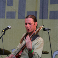 At Positivus 2008, a musician with long hair tied back plays a red bass guitar on stage. He wears a light green shirt and has a focused expression. A microphone is set up in front of him, and a drum set is visible in the background. The stage backdrop features large, partially visible letters, possibly "AB.