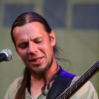 At AB 2008, a man with long hair tied back and a small beard and mustache plays an electric bass guitar on stage. Wearing a beige shirt, he appears focused on his performance, with a microphone in front of him. The background is blurred, indicating stage lighting and possibly a Positivus banner.