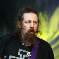 At Positivus 2008, a man with long hair and a beard stands on stage, wearing a black shirt with a white geometric design and a purple guitar strap. He has multiple necklaces, including one with a skull pendant, and appears to be singing or speaking against the dramatic yellow lighting backdrop.