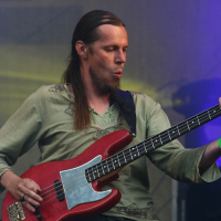A musician with long hair, wearing a light green shirt, plays a red electric bass guitar. He appears to be deeply focused and immersed in the music, with stage lighting in hues of purple and yellow. The setting suggests a live performance at Positivus AB 2008 festival.