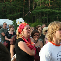 A diverse group of people stands outdoors at a forested location, attending an event reminiscent of Positivus. In the foreground, a woman wearing a colorful headscarf and earrings smiles while looking ahead. Other attendees, casually dressed, are visible in the background among trees and signs with AB 2008 references.