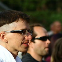 A person with short, dark hair and a shaved section wearing glasses and a white shirt is facing left in an outdoor setting at Positivus 2008. Another person with short dark hair and sunglasses is seen in the background, slightly out of focus. Trees or foliage are blurred in the background.