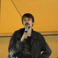 A man with a mustache and glasses is holding a microphone and a piece of paper, speaking under a yellow canopy at the AB 2008 event. He is wearing a dark jacket over a t-shirt with indistinguishable text. His other hand rests on his hip, exuding confidence while Positivus equipment is visible in the background.