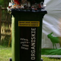 A green trash bin labeled "ORGANISKIE" at the 2008 Positivus festival is overflowing with various waste items such as plastic cups, cans, and paper. Additional litter surrounds the base of the bin. There is a white and green sign in the background and a wooden fence partially visible to the left. The text on the bin reads: "Nemē tī zemi, nemet