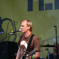 A man is on stage at Positivus, playing an electric guitar and singing into a microphone. He is wearing a brown T-shirt and blue jeans. Behind him, a drummer is partially visible, playing the drums. The background features a large, yellow banner with indistinct black graphics and text, reminiscent of AB 2008.