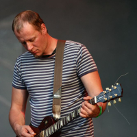 A man with short hair, wearing a striped t-shirt and jeans, is playing a brown electric guitar at the Positivus festival. He stands against a blurred dark background, looking down at the guitar, focused on his performance. His guitar strap is firmly placed on his shoulder.