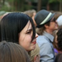 A person with long black hair looks to the side in a crowded outdoor setting at Positivus. They are surrounded by others, including a person in a gray fedora. The background shows blurred trees and people, indicating a possible event or gathering. The person appears thoughtful or pensive.