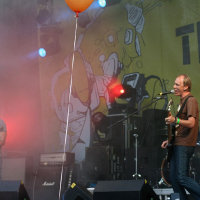 A band performs on an outdoor stage at Positivus. The bassist, wearing a striped shirt, and the vocalist, in a brown shirt, play guitar and sing into a microphone. A drummer is visible in the background. Bright lights, smoke effects, and a yellow backdrop with illustrations and text add to the dynamic scene.