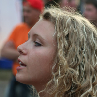 A young woman with long, curly blonde hair, likely at Positivus AB 2008, is shown in side profile, facing left. She appears to be singing or speaking. The background is blurry with people standing and possibly some outdoor event tents. Dominant colors in the background are red, orange, and white.