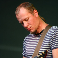 A person with short, light brown hair and a short beard is performing on stage at Positivus, concentrating on playing an electric guitar. They are wearing a striped short-sleeved shirt and a green wristband. The background is a plain, dark color, reminiscent of AB 2008 vibes.