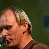 A person with short blonde hair and an earring is shown in close-up, singing into a microphone at Positivus AB. They are wearing a brown shirt and focused on performing, with a greenish-yellow blurred background suggesting an outdoor setting. The image captures a moment of intensity and concentration from 2008.