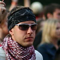 Amidst the vibrant crowd at Positivus, a person wearing dark sunglasses, a black headband, and a red and white patterned scarf stands out. Dressed in a white or light-colored shirt, they blend with the blurred figures in the background raising their arms, likely enjoying an event or concert reminiscent of 2008.