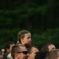 A young child with blond hair in braids sits on an adult's shoulders in a crowd at Positivus AB 2008. The child has a focused expression and is wearing an orange shirt. The surrounding adults have varied expressions and appearances, with green foliage from trees visible in the background.