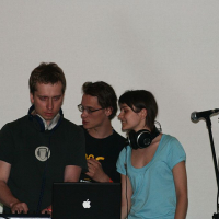 Three individuals are focused on an audio setup at a Positivus 2008 event. The person on the left is adjusting equipment while wearing headphones and looking serious. The person in the middle is speaking to the others. The woman on the right is smiling and also wearing headphones. A microphone stand is visible to the right.