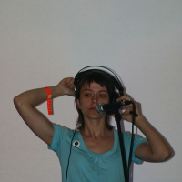 A person with short hair is wearing a light blue shirt with a noticeable AB 2008 badge and a red wristband. They are holding a pair of white headphones with both hands, standing in front of a microphone on a stand against a plain wall background.