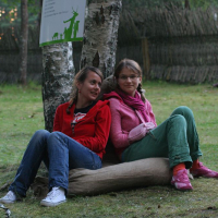 Two young women are sitting on a burlap sack in a grassy outdoor area at Positivus, leaning against a tree. Both are wearing casual clothes; one in a red hoodie, the other in a pink hoodie. They are looking at something off to the side. A few other people are visible in the background, seated near a wooden fence.
