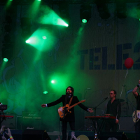 Band performing on a stage with green lighting at Positivus AB, 2008. The lead singer, in the center, holds a guitar and sings into a microphone. Two musicians on either side play keyboards, and one plays a bass guitar. The backdrop displays "TELE2." Balloons float above the partially visible crowd.