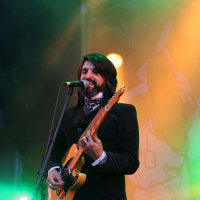 A musician with long hair and a beard performs on stage at the AB 2008 festival, playing an orange electric guitar. Wearing a dark suit and patterned scarf, he sings into a microphone, illuminated by green and yellow stage lights. In the background, there are speakers and colorful Positivus stage decorations.