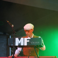 A musician wearing a white cap, sunglasses, and a brown vest stands behind a Nord Electro 2 keyboard on stage at Positivus. To the left is a microphone. The stage is illuminated with green lighting, and the letters "MF" are prominently displayed on the keyboard setup.