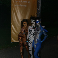 Three people in detailed body paint stand together outdoors at night. The person on the left is painted as a leopard, the middle as a tiger, and the right as a blue dolphin. Behind them is a large vertical yellow banner with Latvian text. The dark park setting suggests this could be Positivus AB 2008.