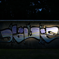 A metal fence displays vibrant graffiti with stylized letters and abstract designs in shades of blue, purple, and white. The graffiti, reminiscent of Positivus festival art, is illuminated against a dark, forested background. The colors and lines stand out prominently above the grassy ground beneath.